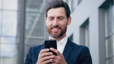 Gülümseyen mutlu adam, elinde telefonla sosyal ağı tarıyor dışarıda duruyor başarılı bir iş adamı iş ya da e-posta mesajlarına cevap veriyor erkek müşteri akıllı telefon kullanarak çevrimiçi dükkandan sipariş veriyor.
