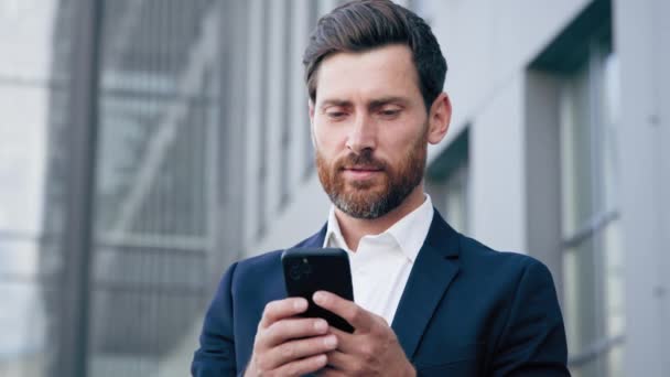 年轻的成功商人手持智能手机站在城市商业中心 用手机写短信 微笑男子经理用手机应用电话远程通信的概念 — 图库视频影像