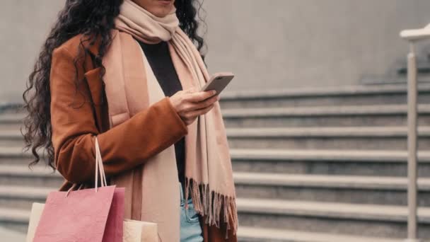 流行のコートで無名の女性買い物客知らない女の子ファッショナブルなスカーフ市内を歩くショッピングバッグパッケージを保持ショッピングバッグ購入チャットブラウジングスマートフォンの携帯電話オンライン割引 — ストック動画