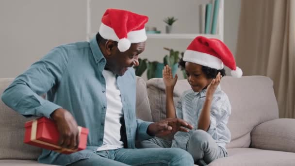 新年を祝う幸せなアフリカ系アメリカ人の家族愛情のある父与え娘クリスマスプレゼントかわいい子供の女の子喜びで驚き子供抱擁お父さん座っている居心地の良い部屋でソファ楽しみ休日 — ストック動画
