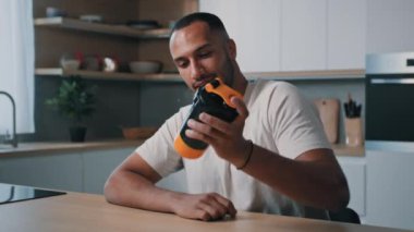 Afro-Amerikalı sporcu erkek sporcu vücut geliştirme uzmanı mutfak masasında oturmuş sıvı kalsiyumla şişe sallıyor protein kokteyli sporlarını karıştırıyor sağlıklı beslenme diyeti yapıyor.