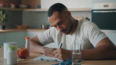 Afro-Amerikalı erkek beslenme uzmanı spor fitness yoga eğitmeni mutfakta oturmuş elmalı vitamin haplarıyla kağıt sayım kalorilerine sağlıklı beslenme diyeti yazıyor.