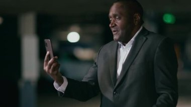 Olgun Afro-Amerikan iş adamı video bağlantısıyla konuşuyor. Cep telefonuyla karanlık odada duruyor. Şaşırmış adam internet konferansında iletişim kuruyor.
