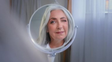 Aynadan yansıyan kadın, yaşlı beyaz kadını şımartıyor. 60 'lı yaşlarda, orta yaşlı, beyaz kadın. 50' li yaşlarda, gülümseyen, buruşuk yüzlü, cilt bakımlı, gri saçlı, nemlendirici.