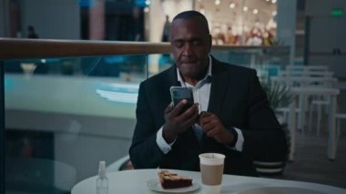 Kafe restoranının kafeteryasında kahve ve pasta dilimleri ile çalışan yaşlı, orta yaşlı bir Afrikalı işadamı. İnternet günlüğü blogunda akıllı telefonları taramak için fotoğraf videosu yapıyor.