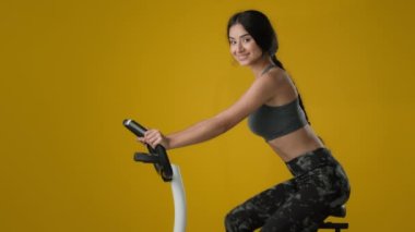 Hint etnik, güçlü, zayıf, sportif kadın, bisiklet süren, bisiklet süren, egzersiz yapan, spor yapan, spor yapan, sarı arka planda spor biniciliği eğitimi alan atletik bayan.