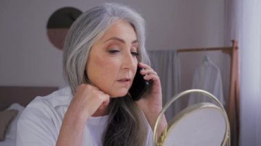 Telefonda konuşan cep telefonu konuşan yaşlı, gri saçlı, 60 'lı yaşlarda kadın aynadaki yansımaya bakan, buruşuk cilde dokunan, uzaktan kumandalı, yaşlı, pürüzsüz cilt nemlendirici bir konuşma.