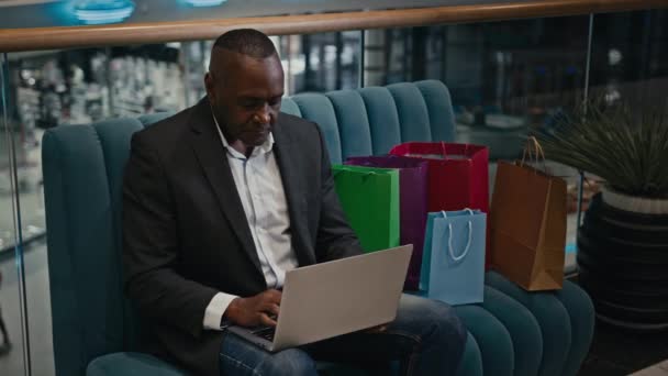 アフリカ系アメリカ人ビジネスマンの労働者の起業家の男性の顧客のショッピングモールのソファに座って袋を購入するラップトップを使用して購入するオンラインストア商品を選択する電子商取引注文を購入 — ストック動画
