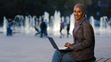 Şehirli fıskiyelerde çalışan, tesettürlü genç müslüman kız tesettüre bakıp mutlu gülümseyen dizüstü bilgisayar uygulaması kullanıyor. Kablosuz bilgisayarı olan etnik kadın.