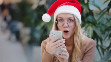 Noel Baba 'nın Noel şapkası ve gözlüklü beyaz kadın Noel Baba' nın mobil zaferine bakıyor. Yeni yıl kutlamaları için indirim teklif ediyorlar.