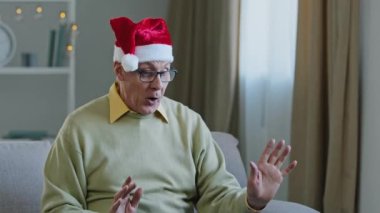Yaşlı Kafkasyalı yetişkin büyükbaba, Noel Baba şapkası takmış evde şarkı söylüyor, dans ediyor, el ele tutuşuyor Noel partisini kutluyor, hediye kutusu sunuyor, video konuşuyor.