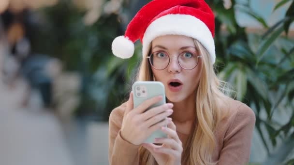 閉じる面白い幸せなショックを受けた白人女性の女の子はサンタクロースのクリスマスの帽子と眼鏡を身に着けている携帯電話の勝利のオファーの達成を見て新年のお祝いの勝利割引 — ストック動画