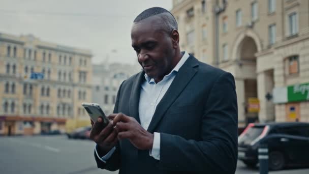 アフリカ系アメリカ人の大人の中年男性の従業員のビジネスマンは 市内の屋外で電話を探して良いニュースを読んで良いニュースを受け取るモバイルメッセージは スマートフォンの成功で幸せなオンライン達成笑顔を提供しています — ストック動画