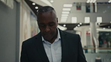 Hareket eden kamera, Afrika kökenli Amerikalı orta yaşlı, yetişkin, 50 'li yaşlarda bir iş adamını alışveriş merkezinin içinde cep telefonuna bakarak cep telefonuyla mesajlaşırken görüntüledi.