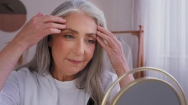 Kapalı Kafkasyalı yaşlı olgun kadın 50 'li yaşlarda aynada yansıyan kadına masaj yapan alna masaj yapan yoga yapan buruşukluk önleyici masaj yapan yaşlılık önleyici cilt bakımı yapan yaşlı kadın.