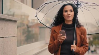 Beyaz, İspanyol asıllı, 30 'lu yaşlardaki bayan iş kadını. Milenyum kadını. Yağmurlu bir sokakta, telefon ve şeffaf şemsiyeyle yürüyor.