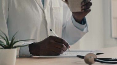 Medikal ceketli erkek doktor klinikte oturur kalem reçetesini not alır Afrikalı uzman doktor pratisyen hekim doktor iş günü çay içerken notlar alır.