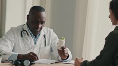 Profesyonel doktor Afrikalı erkek terapist göz doktoru jinekolog kadın hastaya danışmanlık yapıyor. Hastalık kontrolü, sağlık kontrolü. Hasta şişesine antibiyotik ağrı kesici verin.
