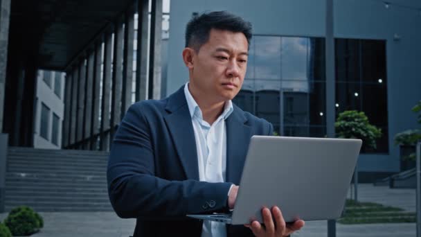 アジア40代の中高年ビジネスマンが都心でノートパソコンの屋外を入力韓国の成人起業家雇用主マンワーカーマネージャー投資家コンピュータで作業オフィス会社の建物の近くに立つ建築家 — ストック動画
