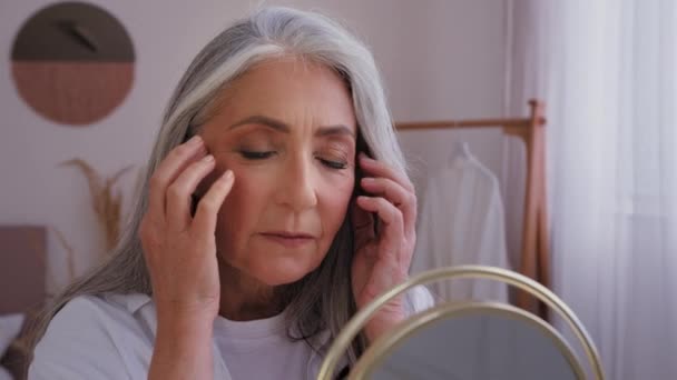 白种人老年女性老年女性50多岁女性看着镜子反射按摩脸抗皱按摩面颊抗衰老提肤护肤运动保湿乳膏 — 图库视频影像