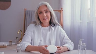 Beyaz, yaşlı, olgun, 50 'li yaşlarda, gri saçlı, vlogger blogcusu evde oturmuş güzellik ürünleri krem kavanozu ve kozmetik etkili video blogu gösteren bir kameraya bakan bir kadın.