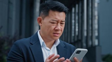 Sinirli, Asyalı, orta yaşlı, lider iş adamı, cep telefonunu açık havada tutuyor. Cep telefonu hata spam mesajından rahatsız. Şehirde yavaş bozuk akıllı telefon bağlantısı sorunu yüzünden üzgün duruyor.