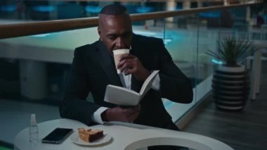 Afrikalı işçi işadamı işveren, kafeterya restoranındaki masada oturuyor, aromatik kahve içiyor, çay içiyor, latte cappuccino içiyor, kitap okuyor, not defteri bilgi metni okuyor.