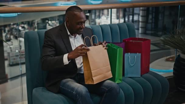 アフリカ系アメリカ人の中高年男性大人のビジネスマンが屋内でモールショッピングセンターに座っているソファの上にショッピングバッグチェック購入バッグギフト購入中を見て楽しむ割引黒金曜日 — ストック動画