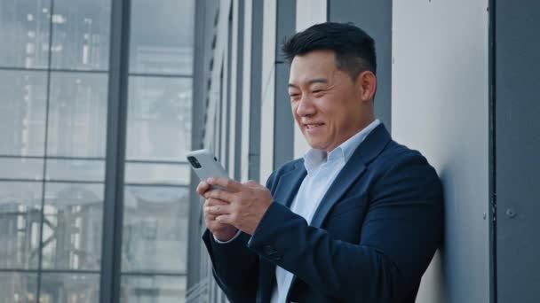 サイドビューアジア40代の中年実業家雇用主男は 市役所企業ビルのガラスの壁の近くに立ってスマートフォンのチャット入力を使用して携帯電話の携帯電話を閲覧 — ストック動画