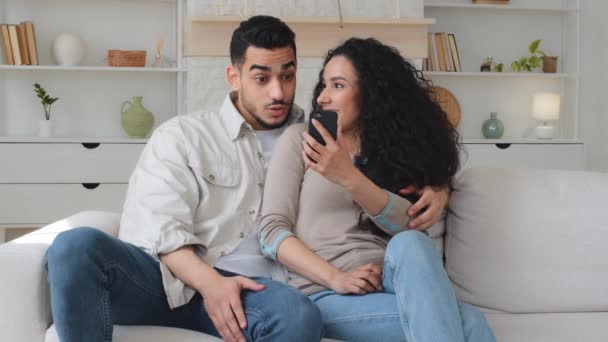 幸せな陽気な女性と男ヒスパニックラティーノ結婚カップルを見てスマートフォン笑顔を話して面白いビデオ写真を見ます上のソファに家のオンライン楽しいモバイルアプリで携帯電話笑いを使用して座って — ストック動画