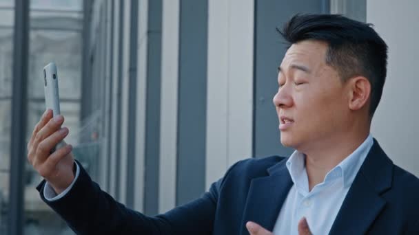 アジアと都市の屋外ビデオ通話韓国の中年の大人と40代のビジネスマン男性インフルエンサー男性雇用主起業家は 携帯電話会議でWebカメラのスマートフォンの仮想チャットオンラインで話す — ストック動画