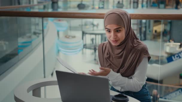 阿拉伯裔穆斯林女商人伊斯兰裔女自由职业者坐在咖啡厅桌旁 在笔记本电脑视频通话商业无线会议在线法律咨询中查阅纸质文件中的数据 — 图库视频影像