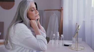 Yaşlı, orta yaşlı, 50 'li yaşlarda, bornozlu beyaz kadın ayna yansımasına bakıyor. Karışık, uzun, parlak saçlarını tarak tarıyor. Alopecia saç dökülme sorunu önleme