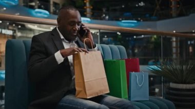 Afrika kökenli, orta yaşlı, yetişkin bir adam alışveriş merkezinin içinde alışveriş merkezindeki koltukta oturmuş, çantalarla konuşuyor cep telefonu konuşmaları, akıllı telefon çekleri, hediye paketleri.