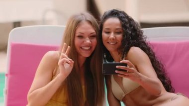 Mutlu kadın moda ünlüleri cep telefonlarında fotoğraf çektiriyorlar havuz kenarında oturup eğleniyorlar. Etnik Latin ve Kafkasyalı arkadaş blogcuların fotoğraf çekimleri komik bir yüz ifadesinin yaz tatilinin tadını çıkardığını gösteriyor