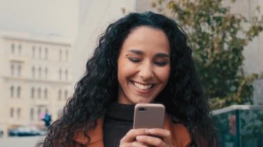 Gülümseyen mutlu İspanyol iş kadını kadın şehir caddesinde cep telefonuyla yürüyen, akıllı telefon takılan, kayan, sohbet eden sosyal medya uygulaması ile mesajlara cevap veren kadın.