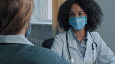 Tıbbi maske takan Afrikalı doktor kadın hasta erkek hastaya boyun ağrısı, sırt kası gerilimi şikâyeti var. Kadın hekim dinle sağlık şikayeti tıbbi danışmanlık yapıyor.