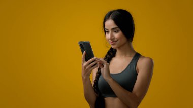Stüdyo sarı arka planda, Hintli 20 'li yaşlarda, spor giyim kuşağında sporcu bir bayan, internet üzerinden yazıcı sohbeti yapıyor, cep telefonuyla spor sohbeti yapıyor.