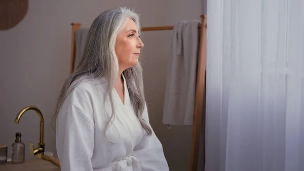 老人60代白人50代の高齢者自宅のバスルームに立って白いバスローブで成熟した豪華な女性 白髪保湿若返りスキンケアと穏やかな祖母の女性 — ストック写真