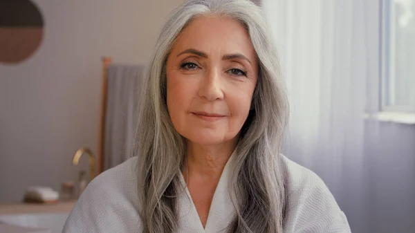 室内の肖像画古い白人50代のシニアモデル白髪熟女60代の女性バスルームにアンチエイジング化粧品の瓶を示しています天然有機日焼け止めミネラルクリーム保湿顔スキンケア — ストック写真