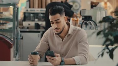 30 'lu yaşlarda Hintli sakallı, etnik kökenli, çok ırklı, işadamı, işadamı, kafeterya masasında oturan, kahve içen, cep telefonu yazıcı, akıllı telefon yazıcı uygulaması kullanan işvereni.