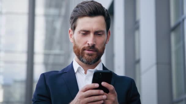 ビジネス管理のためのモバイル電子アプリケーションを使用して バックグラウンドオフィスビルに屋外に立って電話を保持します幸せな男は携帯電話でインターネット上でメッセージを書き込みます — ストック動画