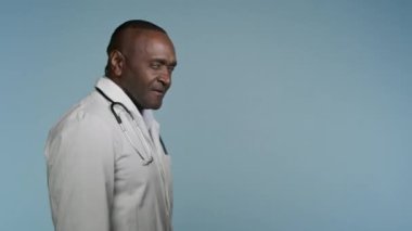 Gri arka plan stüdyosunda yürüyen Afrikalı Amerikalı doktor hastane çalışanı hastaları profesyonel sağlık sigortası el sallamaya davet ediyor.