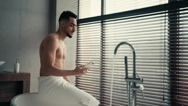 バスルームの衛生で朝風呂インドヒスパニック裸セクシー男座っていますバスタブにタオルで腰を洗う前に保持シャワージェルバームシャンプーのための髪化粧品ボディオイルクリーム男性化粧品 — ストック動画