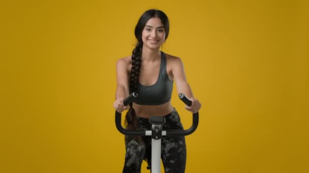 印度苗条快乐的笑容满面的女运动员女运动员女自行车手骑自行车骑自行车锻炼自行车运动器械健身锻炼有氧骑在黄色工作室的大拇指上举手表决 — 图库视频影像