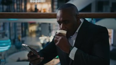 Orta yaşlı Afrikalı girişimci etnik erkek işadamı kafeteryada oturmuş cep telefonuna bakarak Wi-Fi taraması yapıyor akıllı telefonlu internet uygulaması ile telefonla internet işi yapıyor.