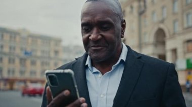 Orta yaşlı Afrikalı girişimci etnik erkek işveren işadamı açık havada cep telefonuna bakan işadamı akıllı telefon internet uygulaması ile çalışan Wi-Fi servis tarayıcısı kullanıyor.