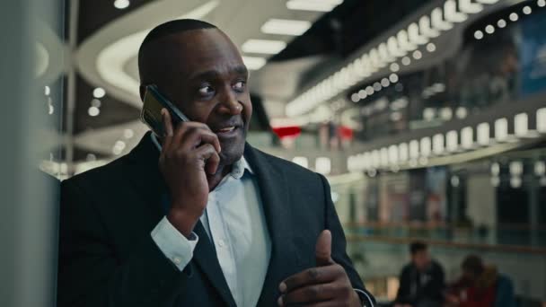 アフリカ系アメリカ人の中年男性の笑顔会社のショッピングモールで携帯電話を話しています 朗らかな大人のビジネスマンが電話を楽しむ幸せな起業家は スマートフォンを呼び出す屋内で笑う話す — ストック動画