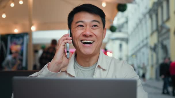 快乐的亚洲男人坐在笔记本电脑旁的咖啡店里 一边用手机聊天 一边快乐的男性自由职业者 一边用智能手机接商务电话 — 图库视频影像
