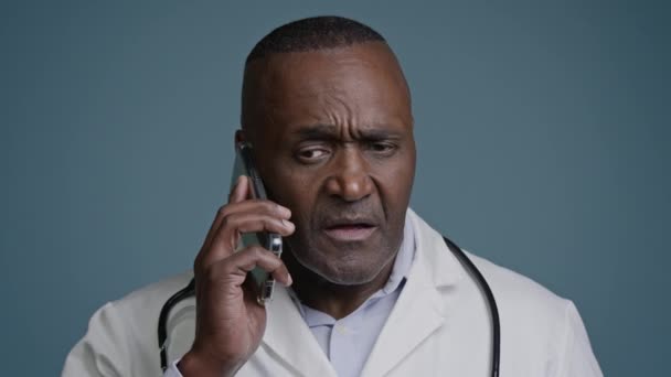 成熟而严肃的医生在电话里与病人交谈不满意的男人医务工作者站在工作室里灰色背景的沟通智能手机远程会诊拒收异议禁令 — 图库视频影像